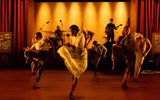 A group of dancers balance on one leg whilst a band plays behind them. The stage is lit in orange light.