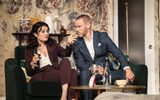 A man and a woman wearing suits sit on a sofa as she drinks a glass of wine.