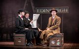 Characters sitting on suitcases look shocked when reading a newspaper. They're sitting in front of a sign reading 'Edinburgh'.