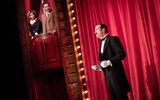 A male wearing a suit stands in front of a red curtain whilst two of the audience peer over a box.