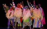 A large group of people wearing brightly coloured clothing stand with their arms raised in the air. Some of them are wearing masks.