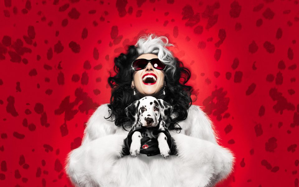 A female with black and white hair wearing a white fur coat. They are holding a dalmatian puppy and laughing.