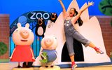 A young female wearing dungarees stretches her arms in the air, she is standing beside Peppa and George in front of the zoo gates.