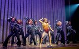 A group of people dance whilst one female sings in a bright sequined outfit.