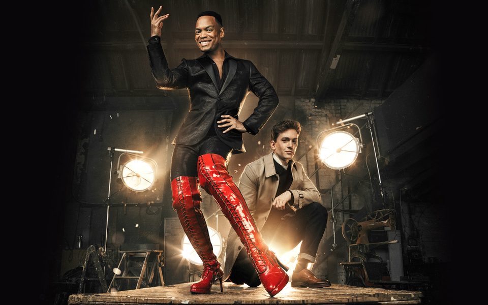 Two males in dark clothing stand on a platform, one is wearing a pair of bright red high heeled boots.