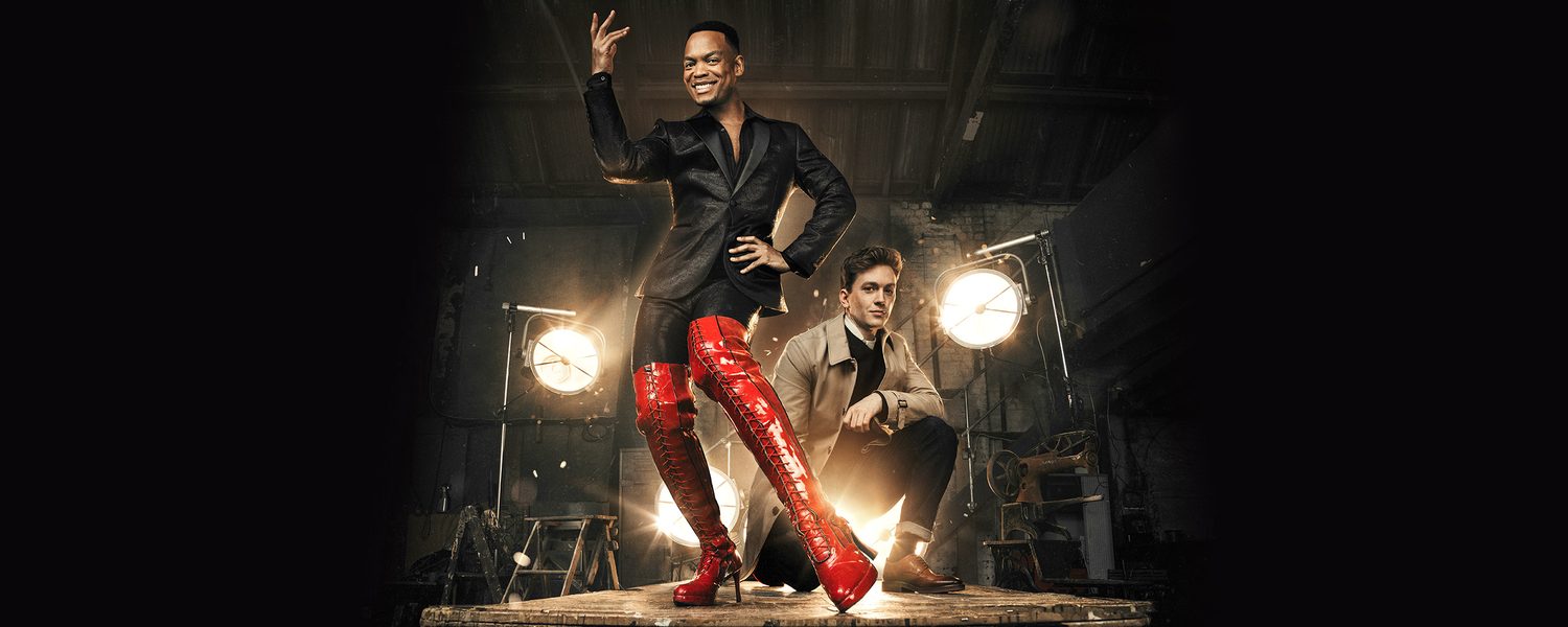 Two males in dark clothing stand on a platform, one is wearing a pair of bright red high heeled boots.