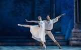 A female balances with one leg extended out behind her, she is wearing a long white dress and is leaning on a male dancer standing next to her.