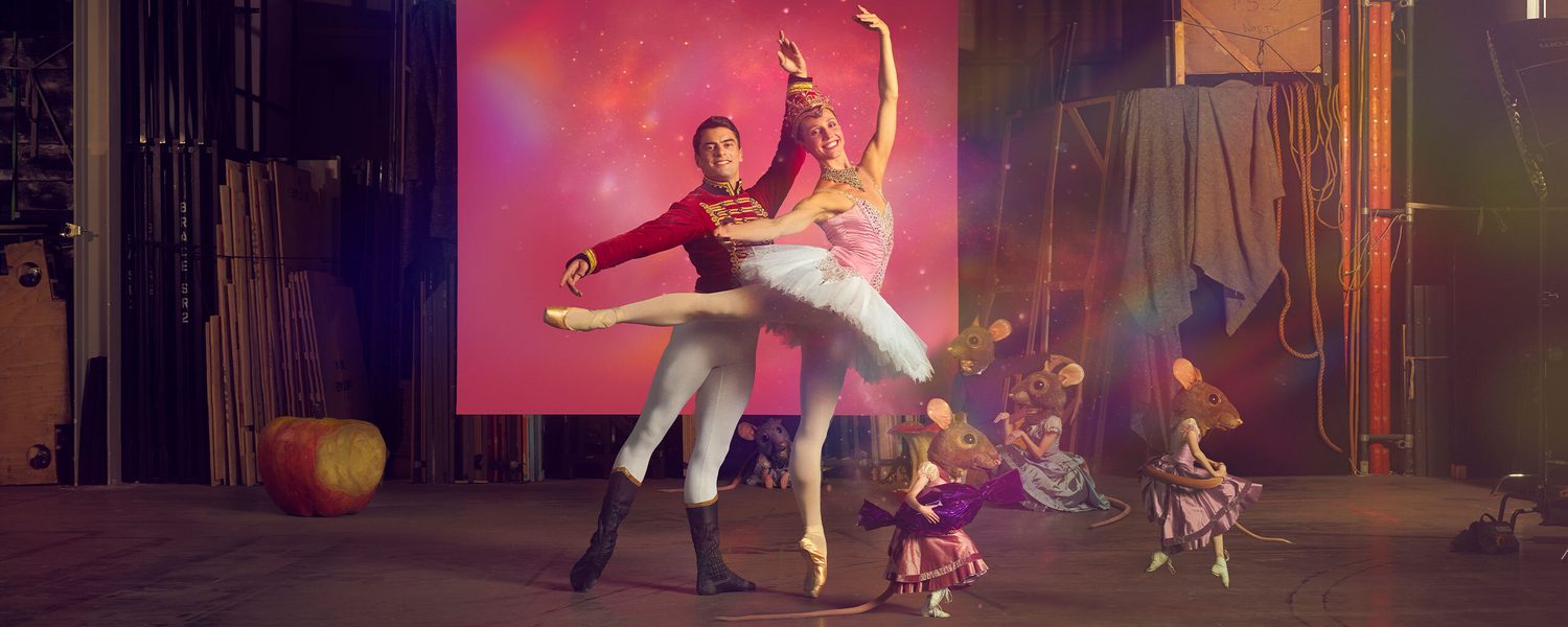 A male and female ballet dancers stand in front of a pink backdrop and there are people dressed in mouse heads beside them.