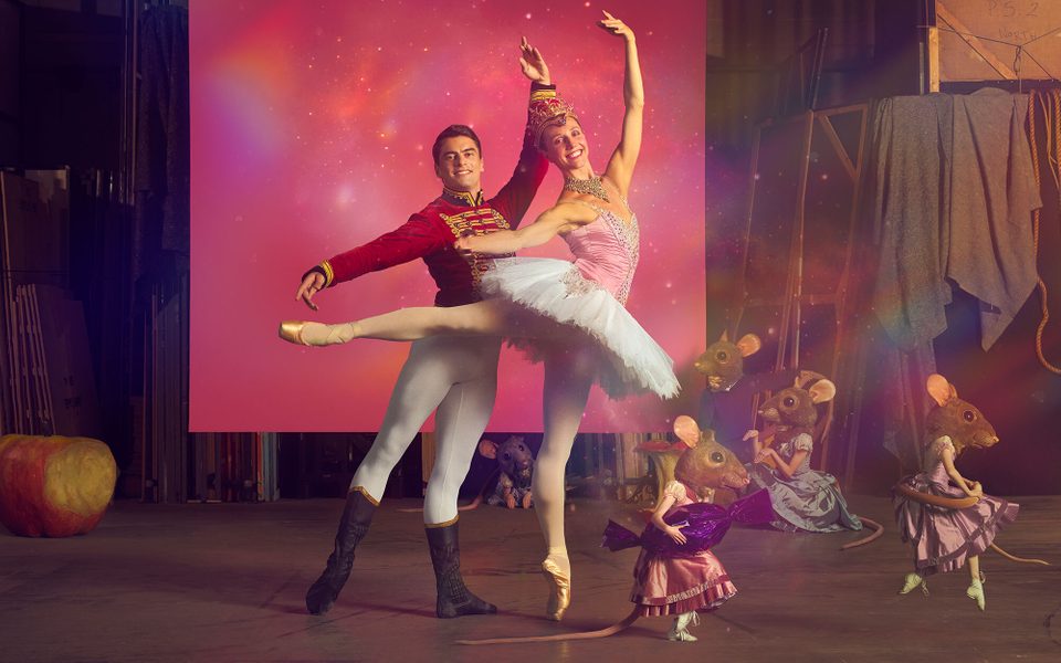 A male and female ballet dancers stand in front of a pink backdrop and there are people dressed in mouse heads beside them.
