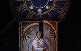 A male steps through a blue and gold wardrobe, there is a large clock above him.