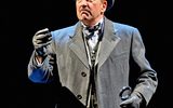 Poirot, a male, wearing a grey suit and overcoat stands in the middle of a dimly lit stage.