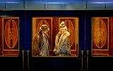 A male and female stand in a train carriage talking. They are both very smartly dressed and are wearing fur coats.