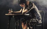 A female sits hunched over a table, she has a cigarette and class of whisky in her hand.