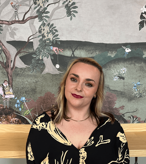 Beth Travers snip A dark haired woman stands proudly in front of scenic wallpaper in green hues featuring intricate drawins to include a black raven, a worn ship's anchor, chorus dancers in black and white stripes