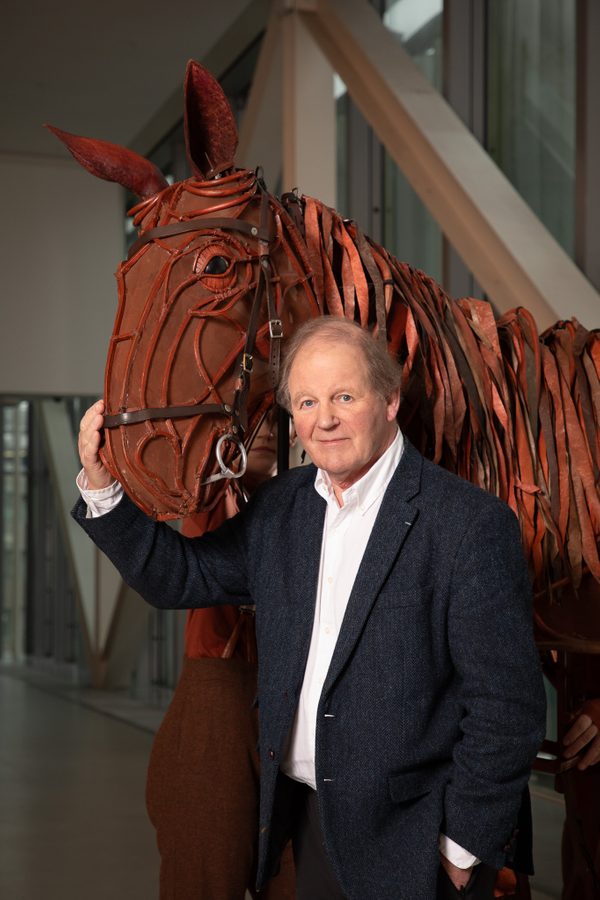 War Horse author Michael Morpurgo dressed in a dark suit and white shirt holding the nose of a conker brown horse puppet depicting Joey the horse.